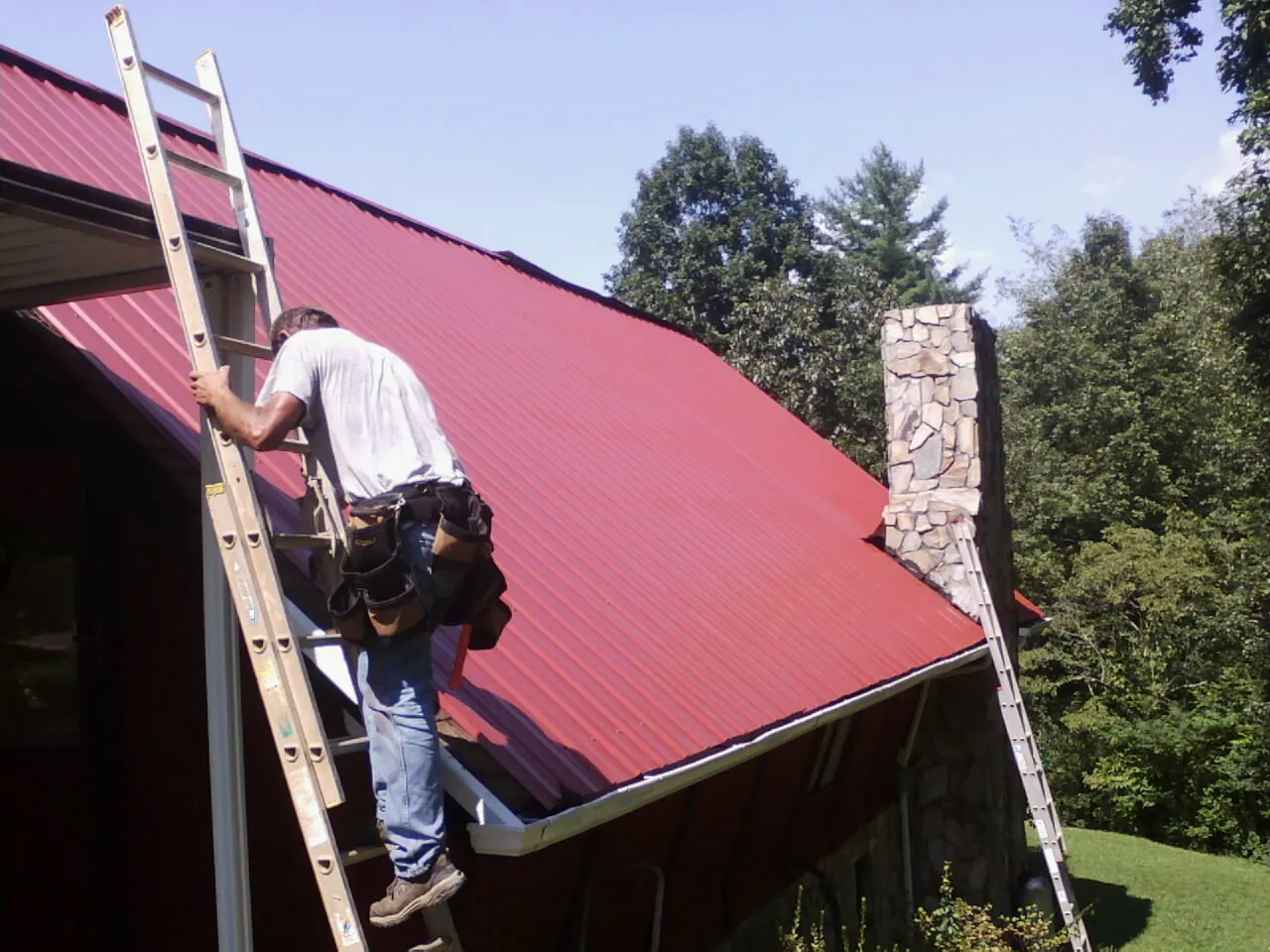 Roofer on a ladder beside a red metal roof and stone chimney — Soffit & Fascia Repair by Roofing Brothers in Mexico